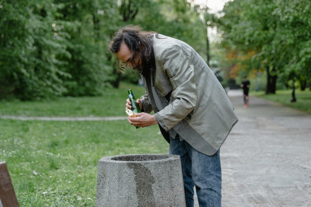 A man searches for food or items in a park trash bin, symbolizing poverty.