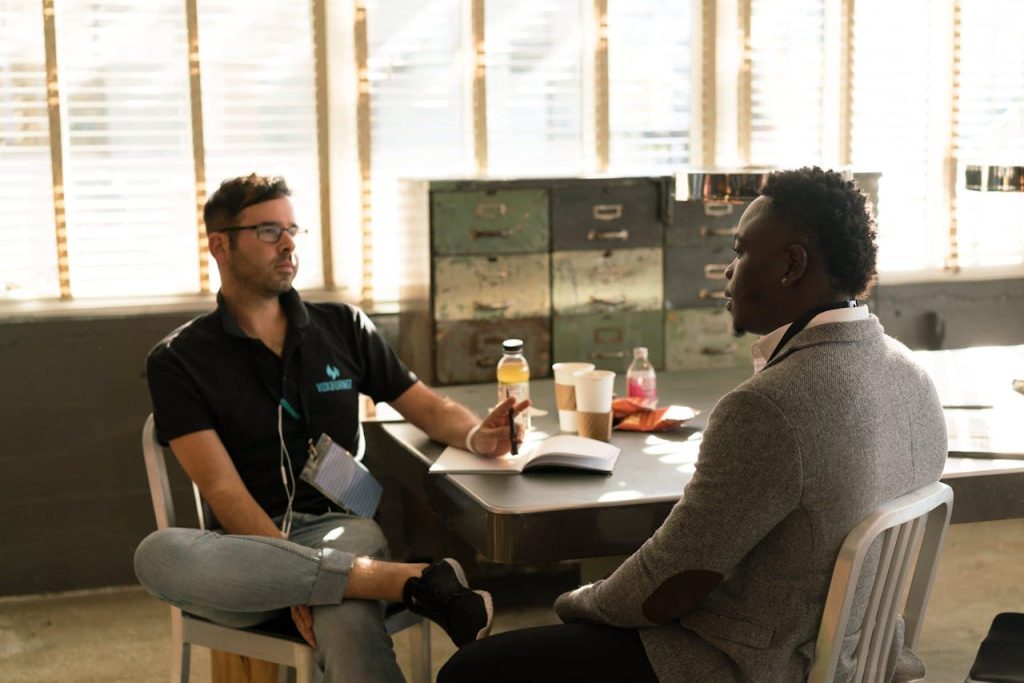 man-wearing-black-polo-shirt-and-gray-pants-sitting-on-white-chair-935977 Two men having a focused business discussion in a sunlit office space.