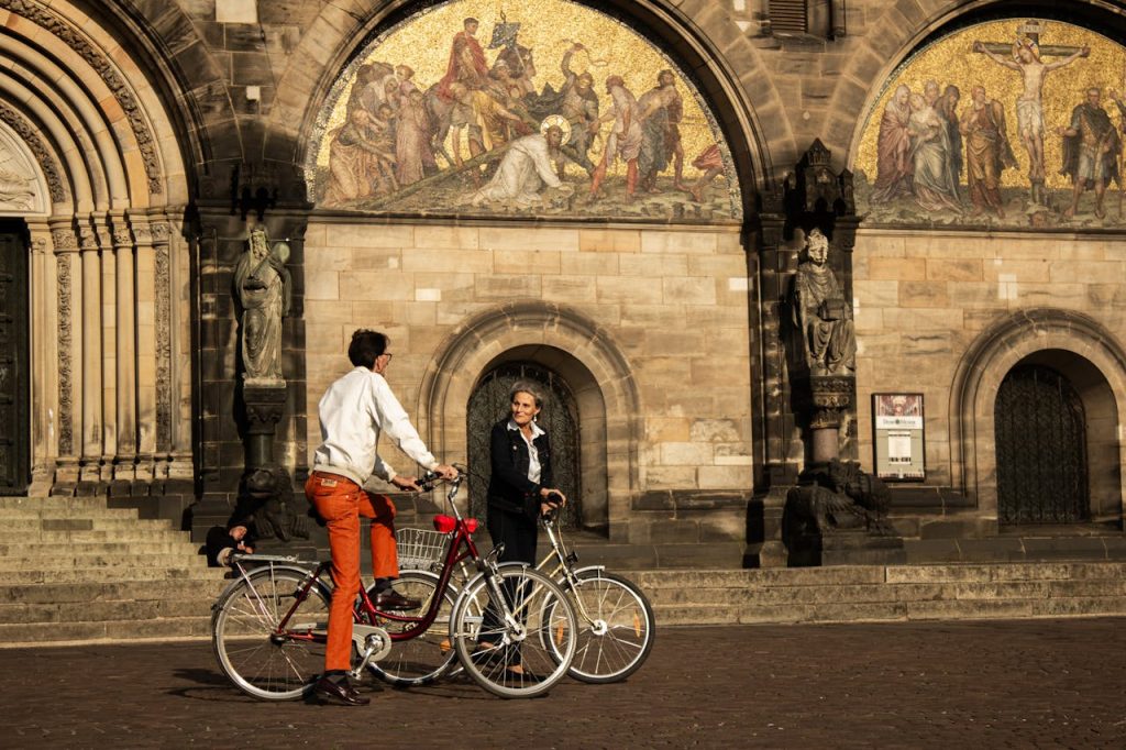 people-riding-on-bikes-in-front-of-cathedral-in-bremen-18722046 Two cyclists ride in front of the ornate arches of Bremen Cathedral, Germany.