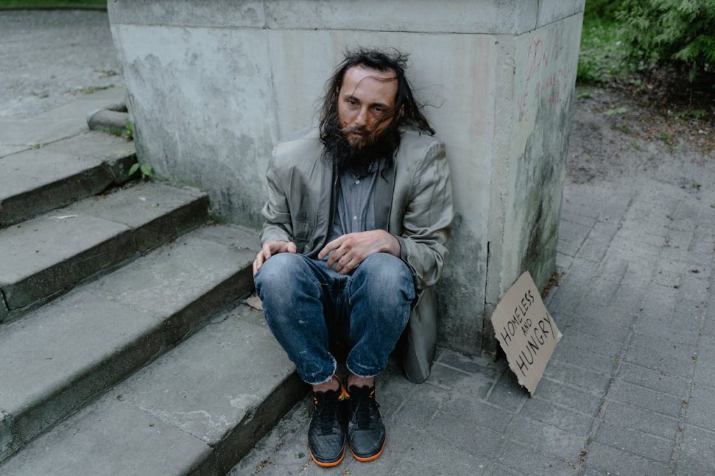 photo-of-a-homeless-man-8078471 A person sitting on steps outdoors with a sign expressing homelessness and hunger.